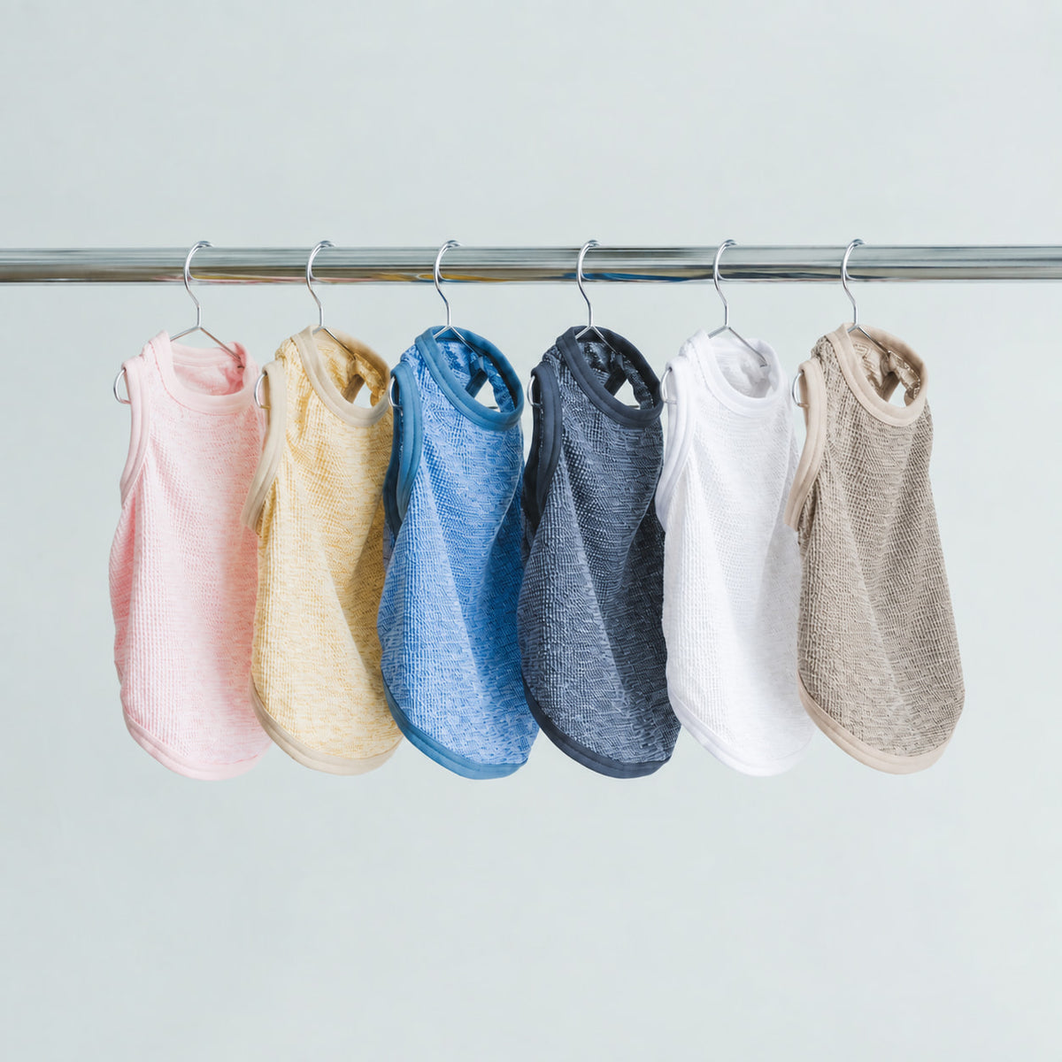ピンク、黄色、青、濃いグレー、白、ベージュの犬用タンクトップ6着がハンガーに並び、縁取りと表面の細かな凹凸が見える。